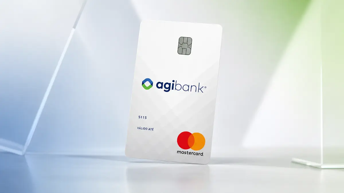 Cartão branco perolado do agibank em pé, com chip, o número 5115 e o logo Mastercard, sobre fundo de vidro azul e verde