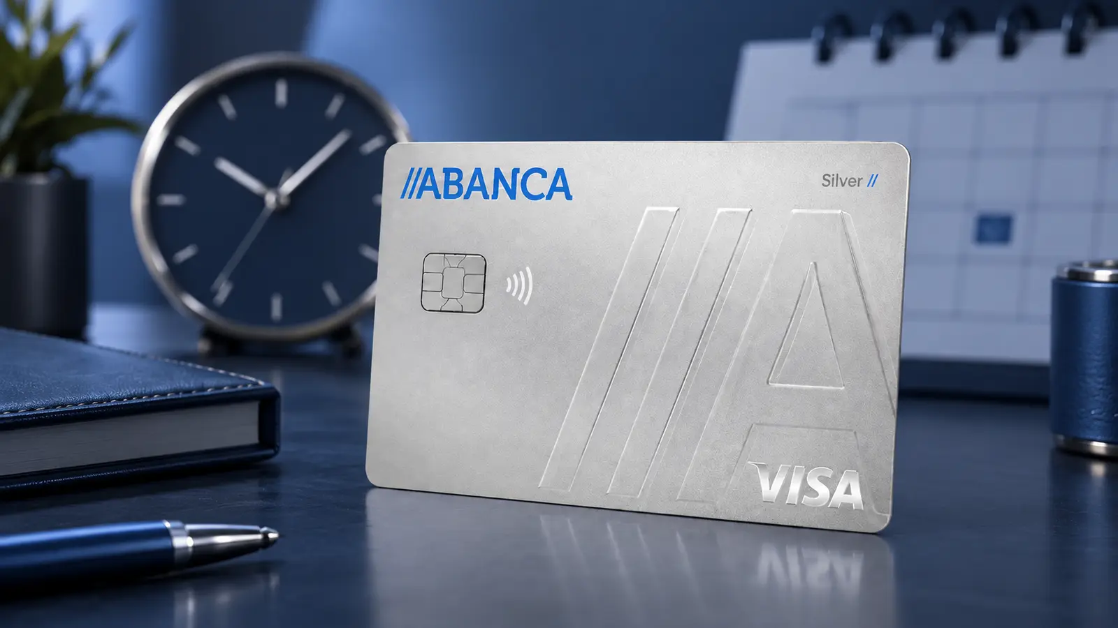 Cartão ABANCA Silver prateado sobre uma secretária azul, diante de um relógio e de um calendário desfocados